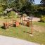 Aire de jeux extérieure 'Trixie' avec structures d’escalade en bois et un bac à sable circulaire dans un parc verdoyant.