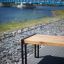 Table moderne en bois Thor long bench avec pieds en métal le long d’un sentier au bord de la rivière avec un pont bleu en arrière-plan