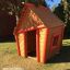 Maison de jeu en bois pour enfants - Crooked House, marron avec bordures rouges, en extérieur.