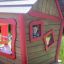 Maison de jeu en bois avec bordures rouges et un enfant souriant regardant par la fenêtre de Anna's Playhouse