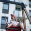 Jeune fille souriante sur une balançoire rouge du 2+2 multiple swing devant un bâtiment moderne.