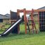 Structure de jeu extérieure en bois Climbing Kiosk Nature avec toboggan, échelle et filet d’escalade dans un jardin