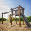 Kampinos wooden playhouse avec toboggans et filet d’escalade sur une aire de jeux sablonneuse.
