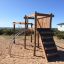 Kampinos wooden playhouse dans une zone extérieure sablonneuse avec des éléments d’escalade et un toboggan.