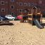 Knight's Castle Nature playhouse with slide dans une aire de jeux sablonneuse avec un immeuble d’appartements en briques