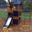 Maison de jeu en bois avec toboggan dans une aire de jeux entourée de feuilles d’automne.