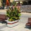 Jardinière Geneva IV avec une végétation verte luxuriante et des fleurs orange sur une place publique pavée entre des éléments en bois