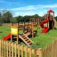 Aire de jeux en bois Mini play avec toboggans, structures d’escalade et balançoires dans un espace herbeux clôturé.