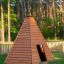 Cabane de jeu en bois de style tipi sur l’herbe avec une forêt en arrière-plan.