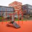 Station de fitness extérieure New Age Fit MEDIUM avec des structures orange devant un bâtiment moderne.