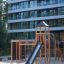 Structure de jeu en bois Explorer castle with slide sur une zone sablonneuse devant un bâtiment moderne