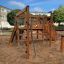 Kiosk Modern Nature wooden playhouse sur une aire de jeux sablonneuse entourée de bâtiments et d’arbres.