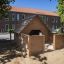Maison de jeu en bois Veranda House Nature dans un bac à sable avec des arbres et un bâtiment en arrière-plan