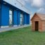 Structure en bois Nature Playhouse sur une pelouse herbeuse devant un bâtiment moderne bleu.