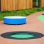 Round trampoline (100 cm) intégré dans une surface d’aire de jeux avec tapis de saut vert et bordure noire.