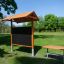Salle de classe en plein air avec bancs en bois, toit pare-soleil, tableau noir et pots de fleurs dans un espace vert extérieur
